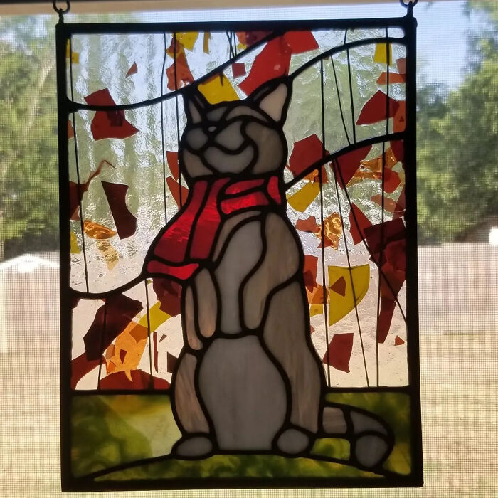 Stained glass art of a cat wearing a red scarf, a vivid pop culture icon in colorful stained glass signs hanging by a window.