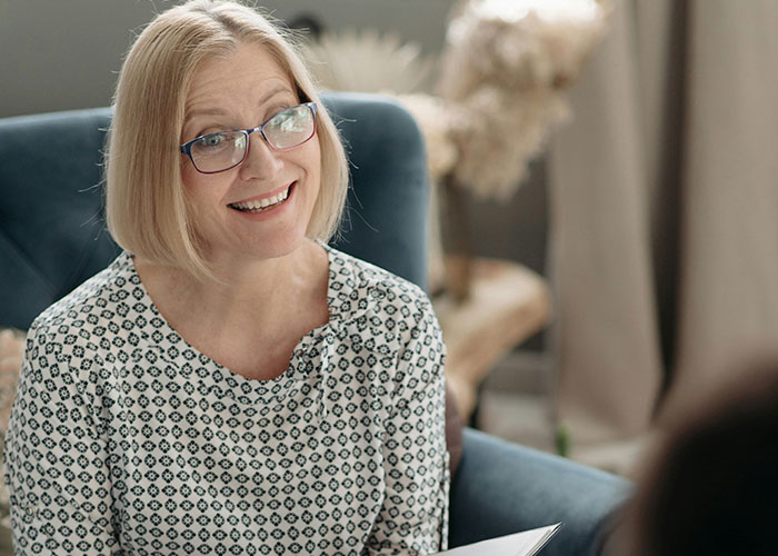 Smiling middle-aged woman with glasses talking in a home setting, depicting shocked reactions to unhinged things their MIL said.