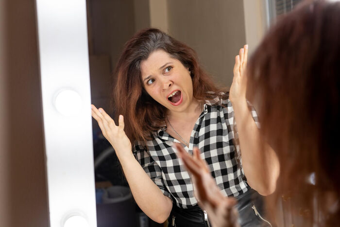Woman in checkered shirt making shocked face in front of mirror, illustrating bizarre things seen through neighbor’s window.