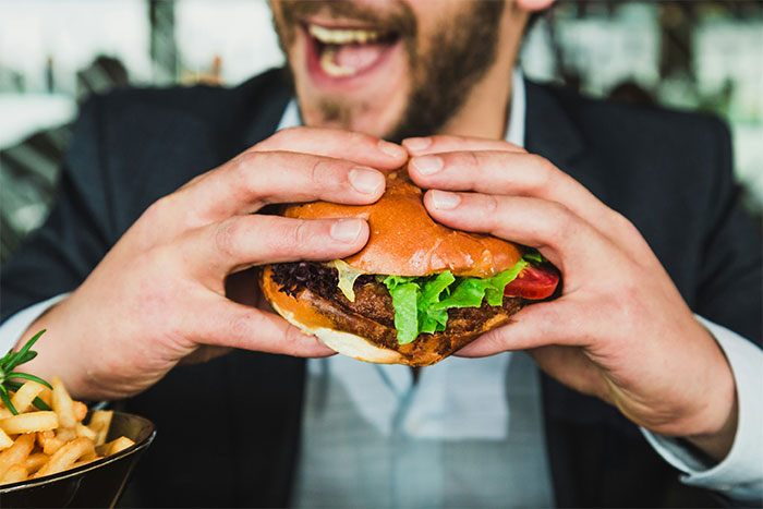 Man in business attire holding a juicy beef burger with fries nearby, illustrating employee pushback against vegan-only club. Man in business attire holding a juicy beef burger with fries nearby, illustrating employee pushback against vegan-only club.