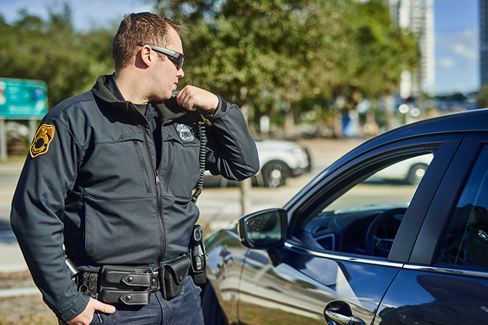 Police officer talking into radio next to a car in daylight, related to half-brother arrested for stealing vehicle case