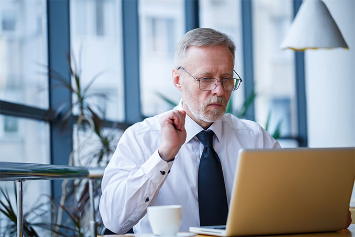 Man in office wearing glasses and tie, working on laptop while reacting to loud Christmas music nearby.