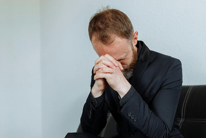 Man in black suit sitting with head down and hands clasped, depicting the boss’s hire crush situation going wrong. Man in black suit sitting with head down and hands clasped, depicting the boss’s hire crush situation going wrong.