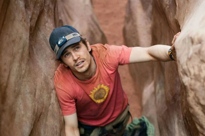 James Franco wearing a cap and red shirt, climbing between narrow rock walls in an outdoor setting.