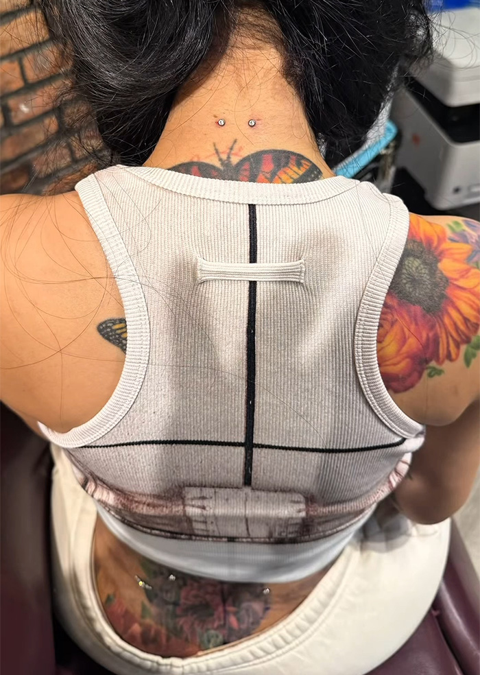 Close-up of neck and back piercings with colorful butterfly and flower tattoos on skin beneath white tank top. Close-up of neck and back piercings with colorful butterfly and flower tattoos on skin beneath white tank top.