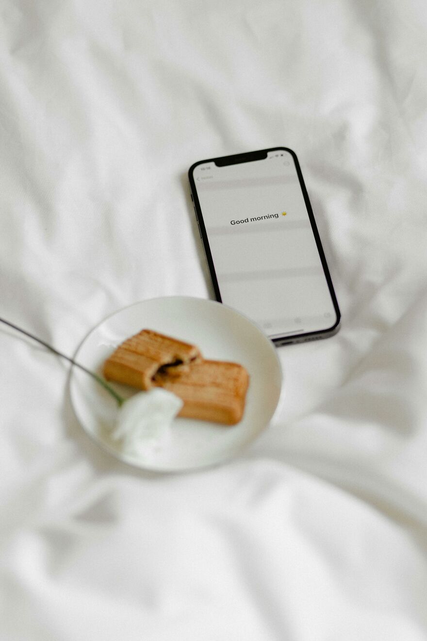 Smartphone displaying a good morning message on a white bed next to a plate with a pastry and a white flower. Smartphone displaying a good morning message on a white bed next to a plate with a pastry and a white flower.