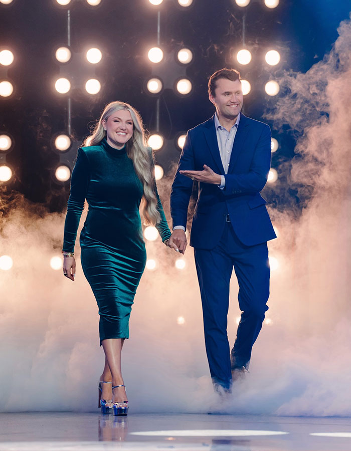 Erika Kirk and husband Charlie walking hand in hand on a foggy stage with bright lights in the background. Erika Kirk and husband Charlie walking hand in hand on a foggy stage with bright lights in the background.