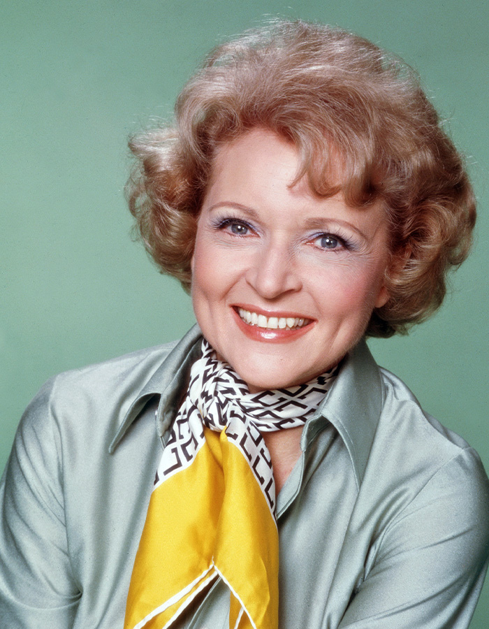 Smiling woman with curly hair wearing a patterned scarf and light green shirt, representing 55YO celebs from different eras.