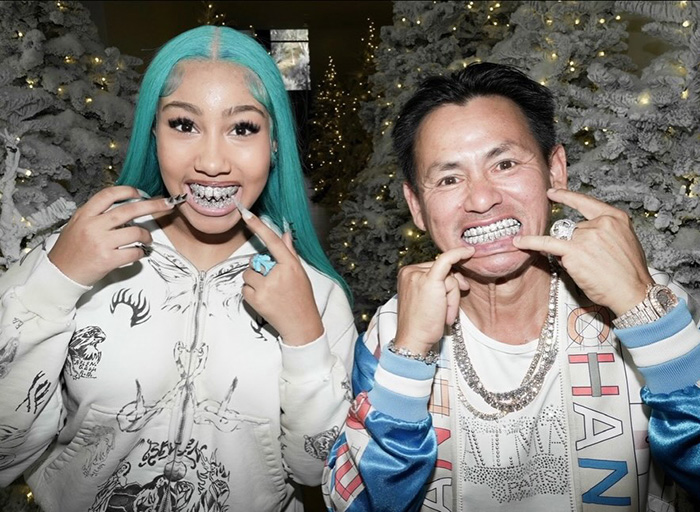 Two people showing off diamond grills, posing in front of snow-covered trees with holiday lights, trendy streetwear style.