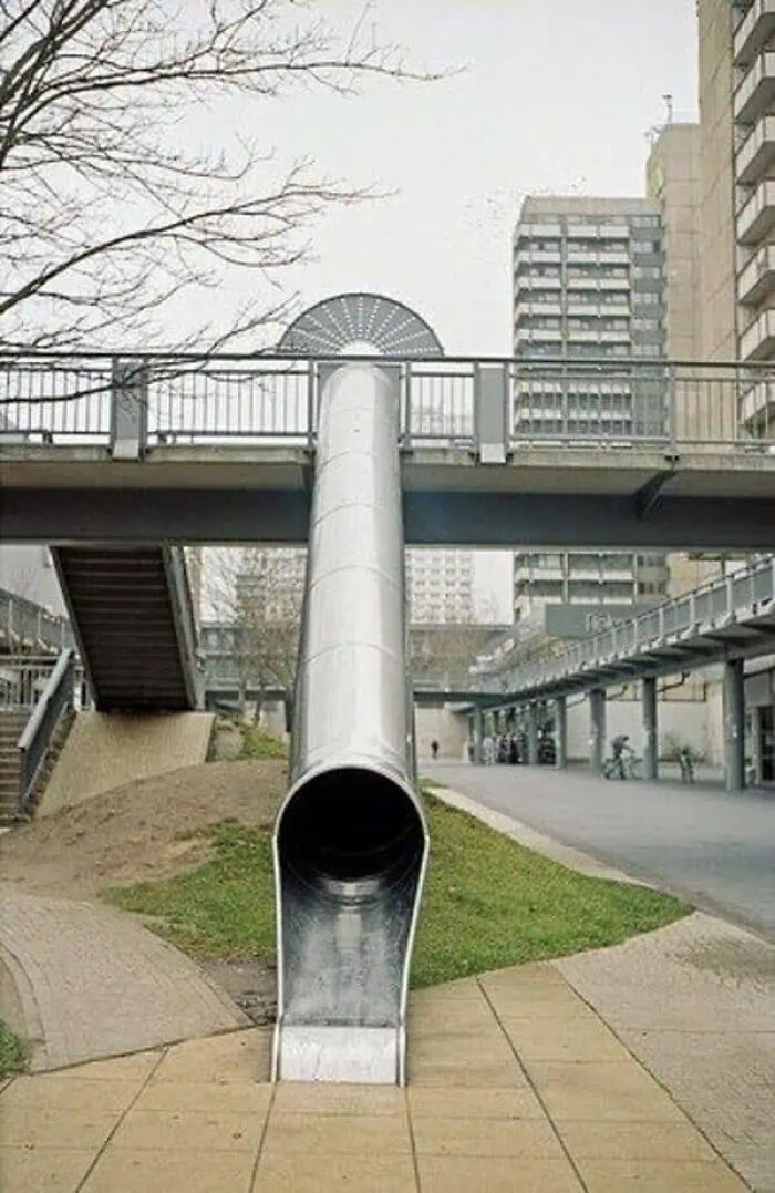 A trippy slide descending from a pedestrian bridge in an urban area, blending architectural elements and unusual design.