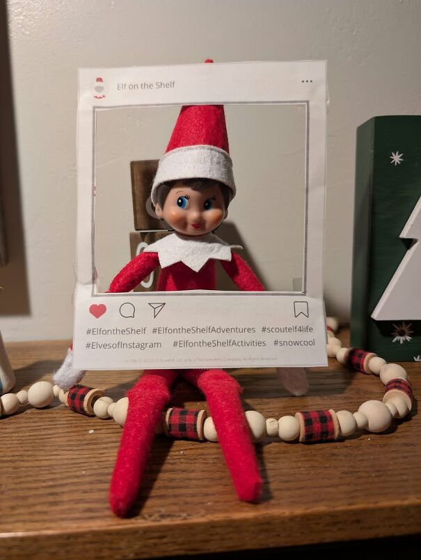 Elf on the Shelf doll holding a paper frame mimicking social media with festive wooden bead garland on a table.