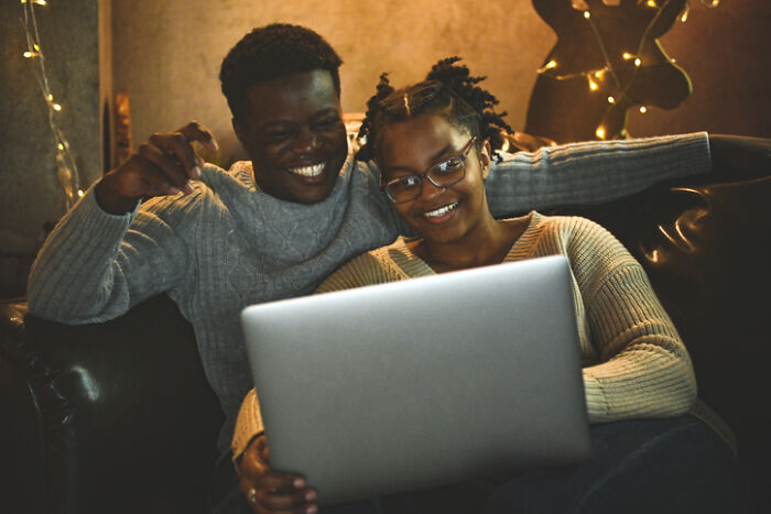 Couple enjoying at-home date night ideas on a laptop, sitting cozy on a couch with warm lighting and string lights.