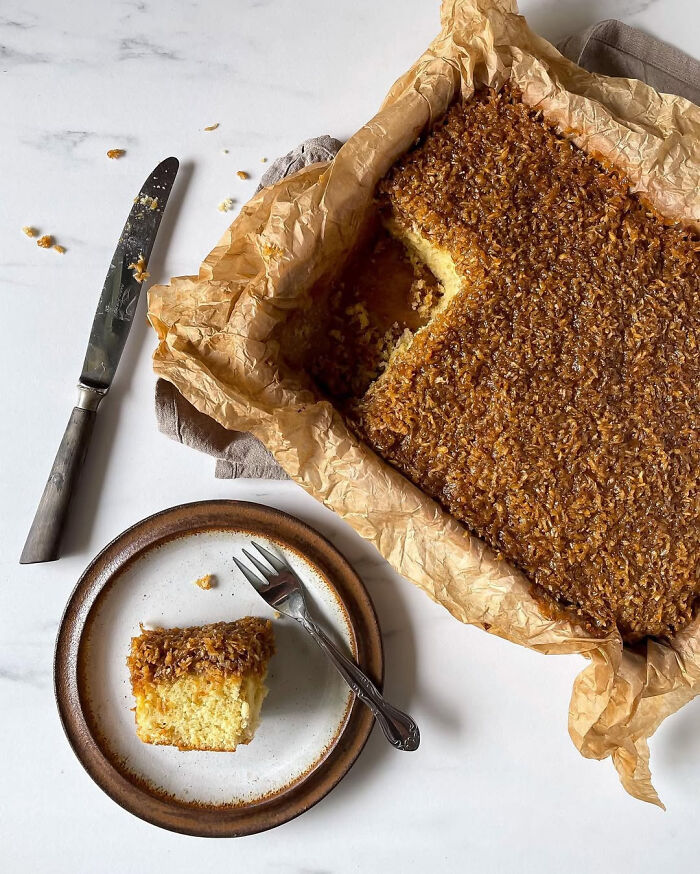 Slice of homemade Christmas dessert with a coconut topping on a plate next to a baking tray with more dessert.