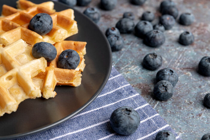 Waffles topped with blueberries on a black plate, surrounded by scattered fresh blueberries on a textured surface.