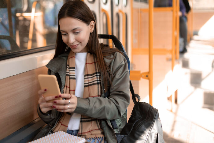 Young woman on public transport smiling at her phone, capturing embarrassing moments people do when unobserved.