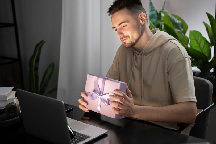 Young man holding a wrapped gift, smiling at laptop screen inside a cozy room, representing crazy things to make an ex pine.