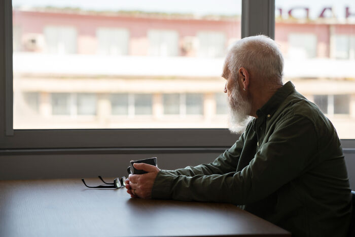 Elderly man with a beard sitting by a window holding a cup, reflecting, related to secrets about celebrities shared by insiders.