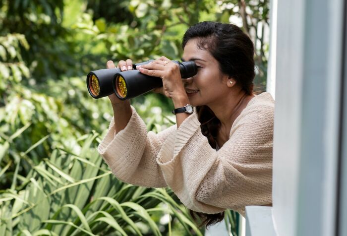 Woman using binoculars outdoors, curious and watching, illustrating bizarre things seen through a neighbor’s window.