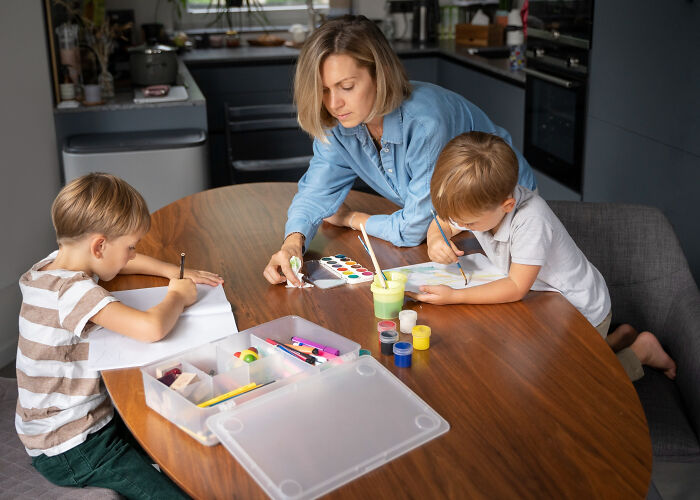 Woman helping two children with painting and drawing at a wooden table using small tricks that make being a grown-up easier.