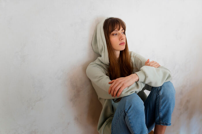Young woman in hoodie sitting against a wall, looking thoughtful and highlighting family drama emotions.