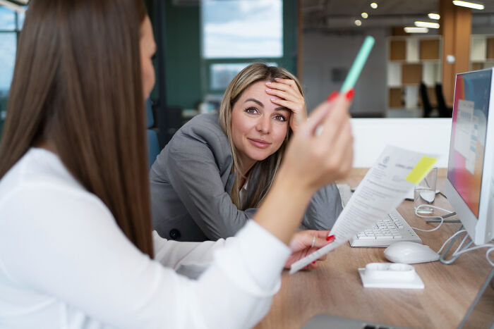 Two female coworkers in an office, one frustrated and the other holding a pen, illustrating infuriating work mindset.