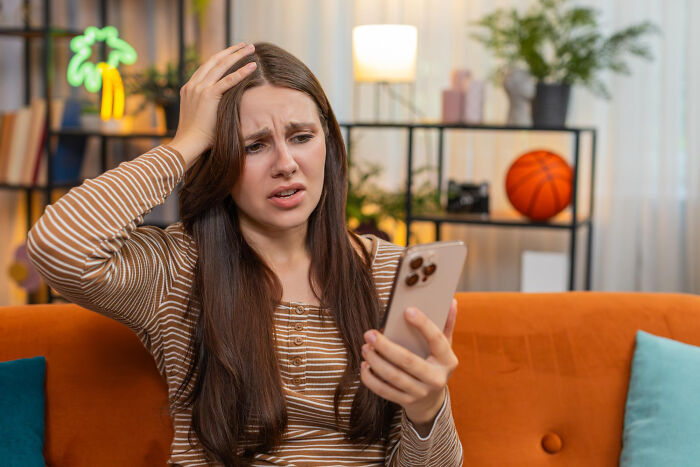 Young woman looking confused at her smartphone while sitting on an orange couch illustrating small tricks for being a grown-up easier.