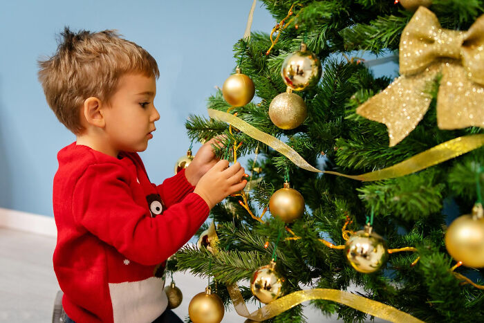 Young boy in a red sweater carefully decorating a Christmas tree, creating a strange and specific kid's insult moment.