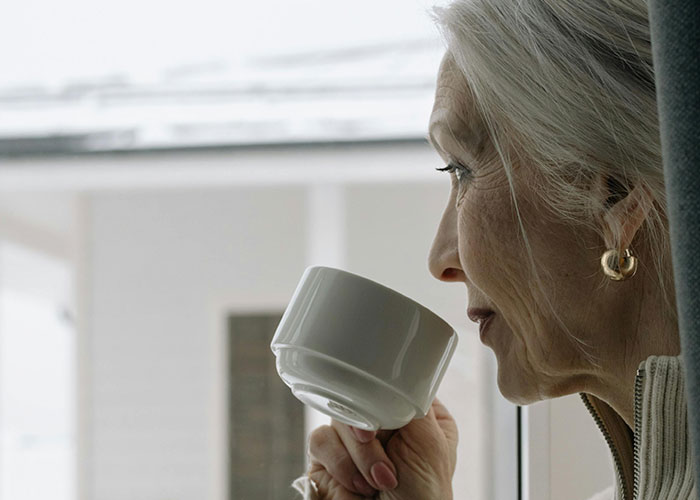 Older woman sipping tea by window, reflecting on shocking unhinged things her MIL just said to her.