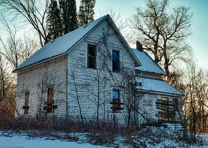 Abandoned house with boarded windows surrounded by leafless trees in a remote and isolated place with snow on the ground.