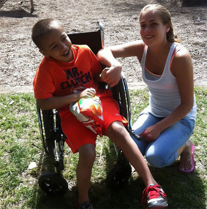 Woman kneeling beside smiling boy in wheelchair outdoors, representing Cinnabon worker’s daughter reacting to racist tirade. Woman kneeling beside smiling boy in wheelchair outdoors, representing Cinnabon worker’s daughter reacting to racist tirade.