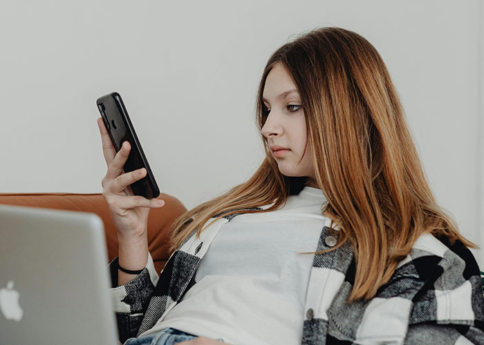 Young woman reading insane things her ex did on phone while relaxing on couch with laptop nearby at home.