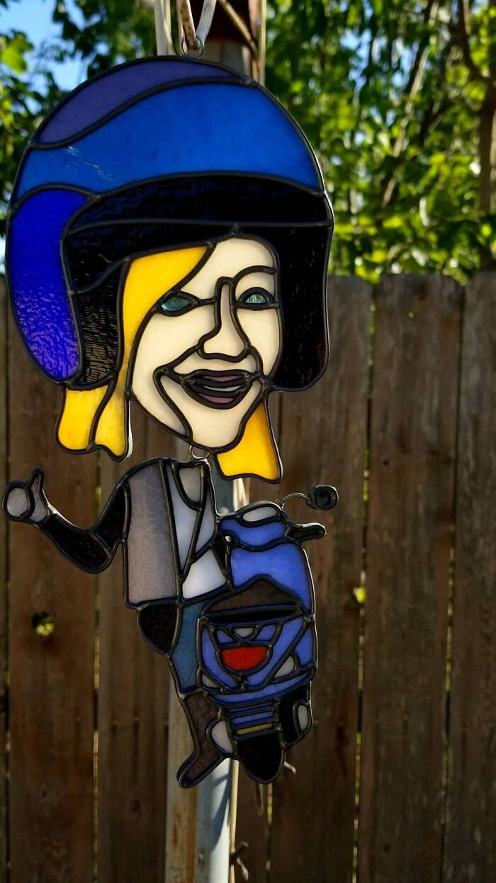 Vivid stained glass sign of a person with blonde hair wearing a blue helmet and riding a blue scooter outdoors.