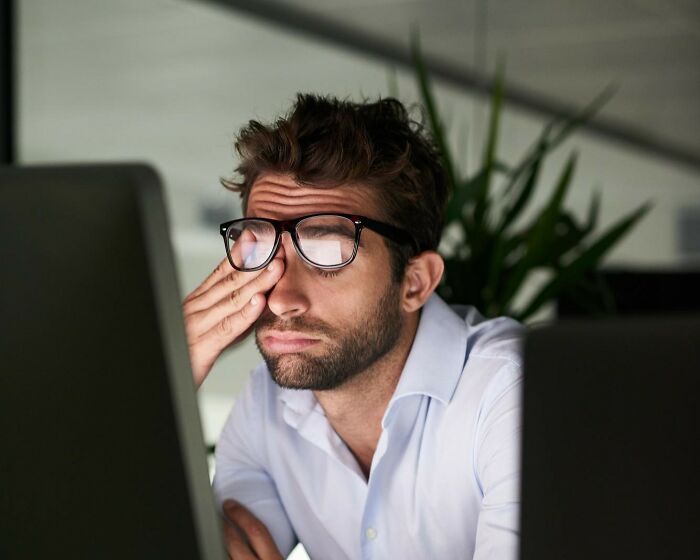 Tired man in glasses at computer rubbing his eyes, illustrating infuriating work frustrations with old habits.