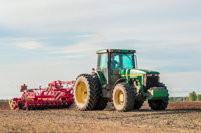Green tractor with red farming equipment working in a field illustrating infuriating examples of we’ve always done it this way at work.