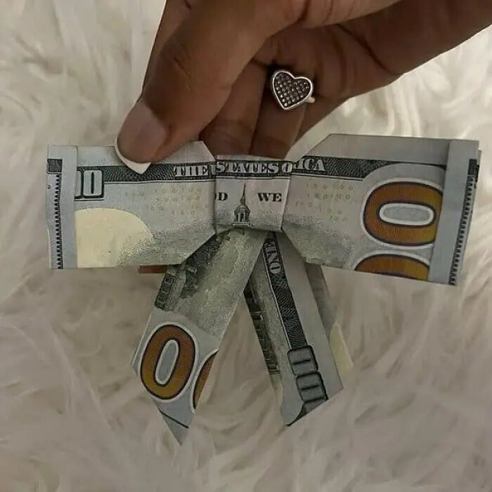 Hand holding a trippy folded hundred-dollar bill shaped like a bow tie against a soft textured background.