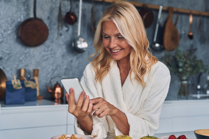 Woman in a white robe smiling at her phone in kitchen, illustrating crazy things people do to make their ex pine for them.