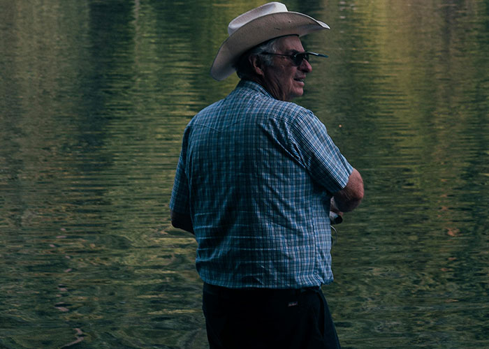 Older man wearing a cowboy hat fishing alone by a calm lake, capturing creepy and mysterious moments in remote places