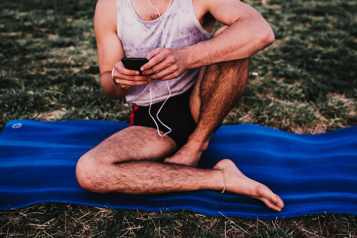 Young man sitting cross-legged on a blue mat outdoors, using a smartphone and wearing earphones during a break.