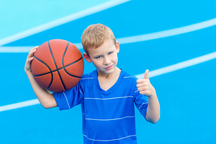 Young boy holding a basketball on court, giving thumbs up, capturing a kid's strange and specific insult moment.