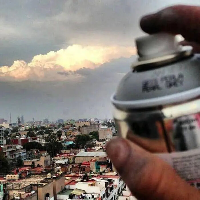 Hand holding spray can creating trippy picture effect with cityscape and dramatic clouds in the background.