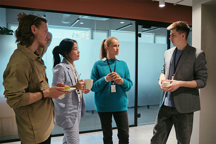 Four employees in a modern office discussing as one holds a plate with an apple and another with a cup of coffee.