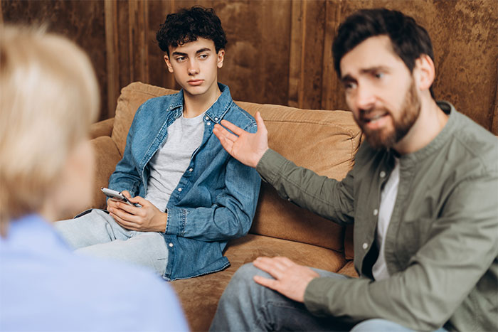 Teen sitting tense on couch while parents argue, highlighting fear and family conflict involving violent sister.