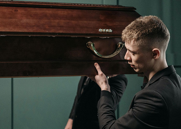 Young man in formalwear carrying a wooden coffin, illustrating people who work in luxury industries and rip-offs.