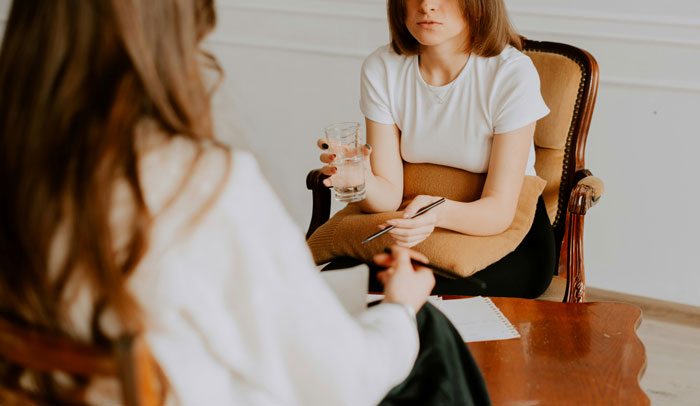 Two women in a serious conversation, one holding a glass of water, depicting next-level evil kids feared by people.