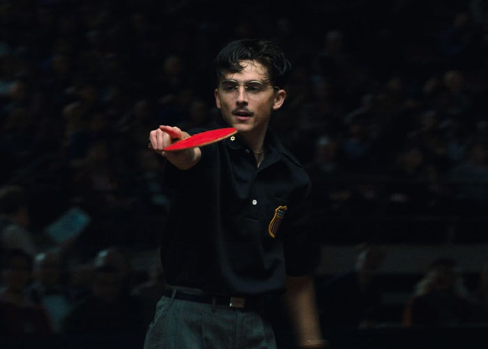 Timoth&eacute;e Chalamet playing ping pong wearing glasses and a black shirt in a dimly lit setting.