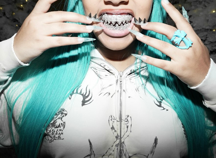 Person with long turquoise hair and spiked grill, wearing white hoodie and displaying long nails and rings in close-up photo.