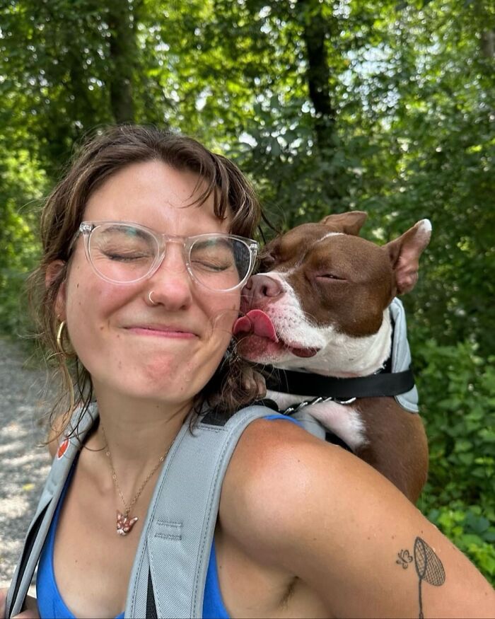 Woman smiling with a dog in a backpack licking her face during an outdoor city adventure among green trees.