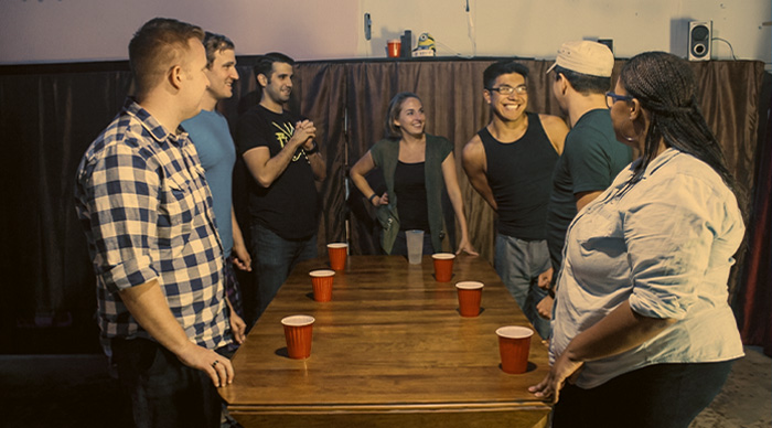A group of friends playing the best party game of all time with red cups around a wooden table indoors.