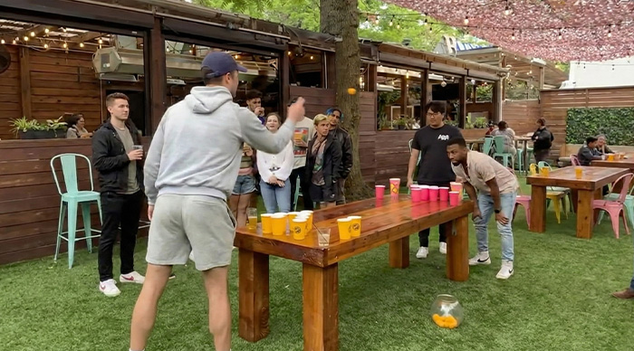 Group of friends playing the best party game outdoors with cups on a wooden table in a lively backyard setting.