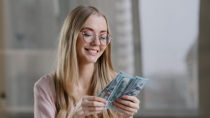 Young woman with glasses smiling while counting money, unaware of potential big family drama unfolding around her.